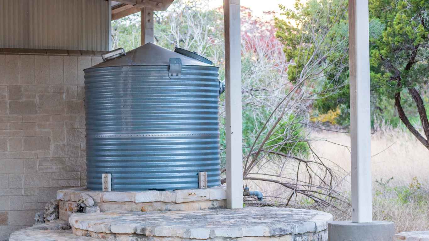 Corrugated steel water storage tank on top of flagstone masonry structure, with small stone fountain in front
